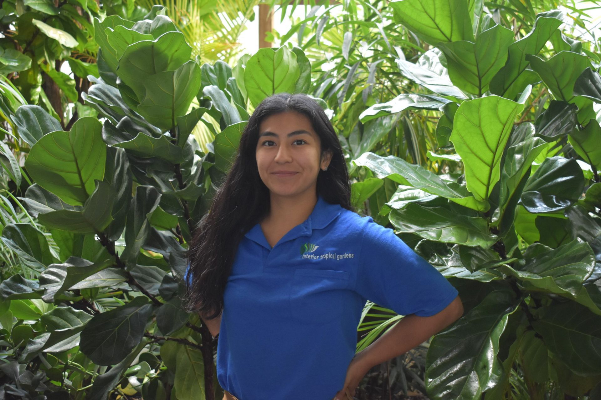 Woman in blue shirt poses in front of large green plants.
