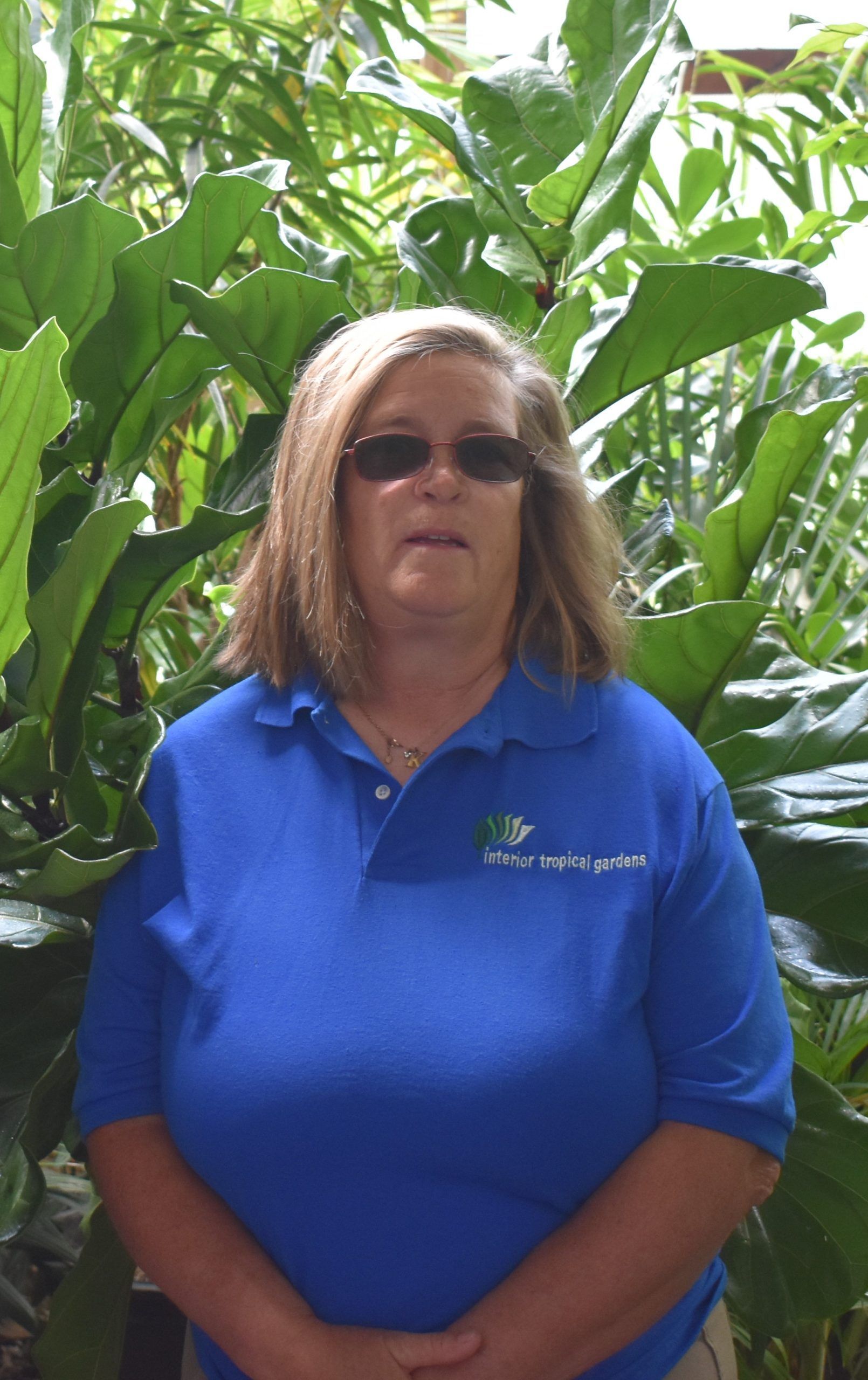 Woman wearing sunglasses and a blue shirt stands in front of green plants.