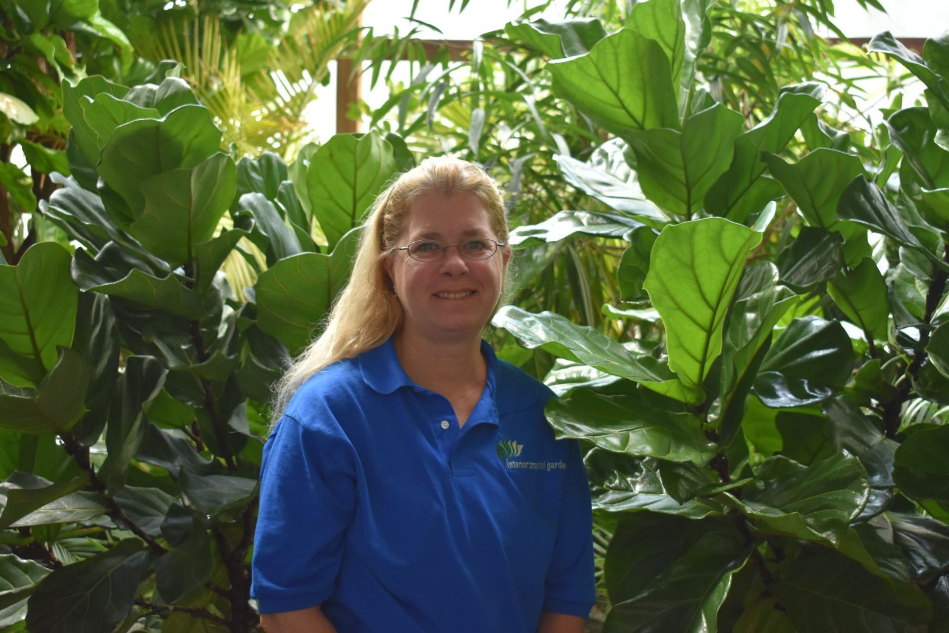 Woman in blue shirt smiles in front of large green-leafed plants.