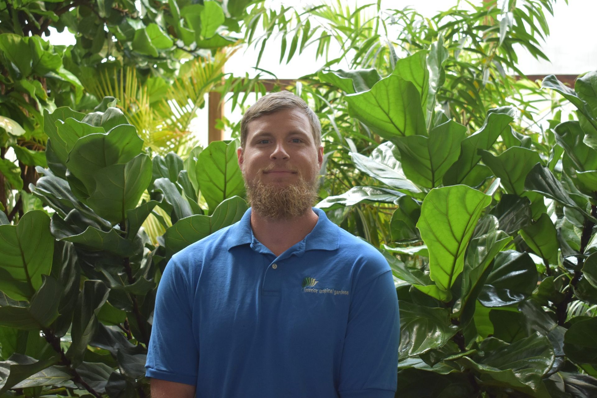 Man in blue shirt, light beard, stands in front of lush green plants.