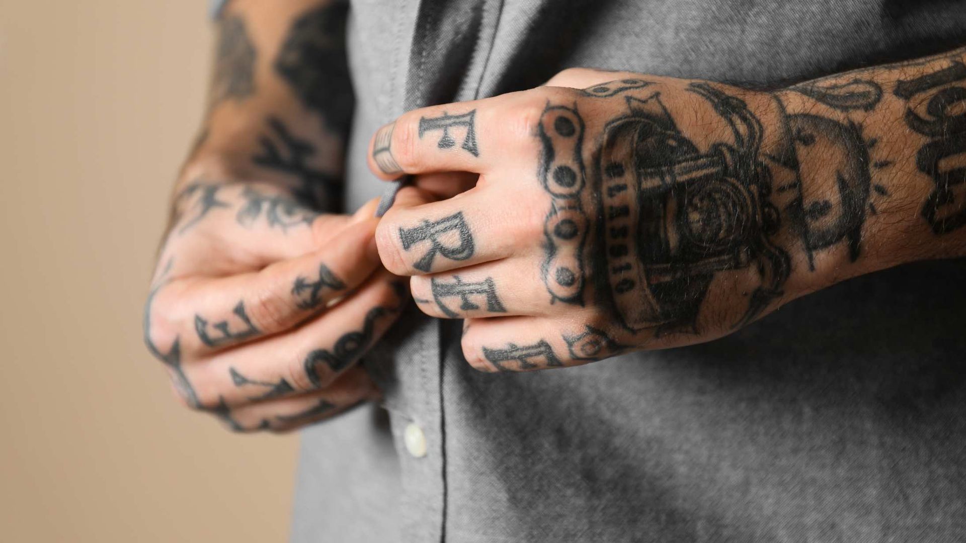 Close-up of heavily tattooed hands buttoning a grey shirt against a neutral background.