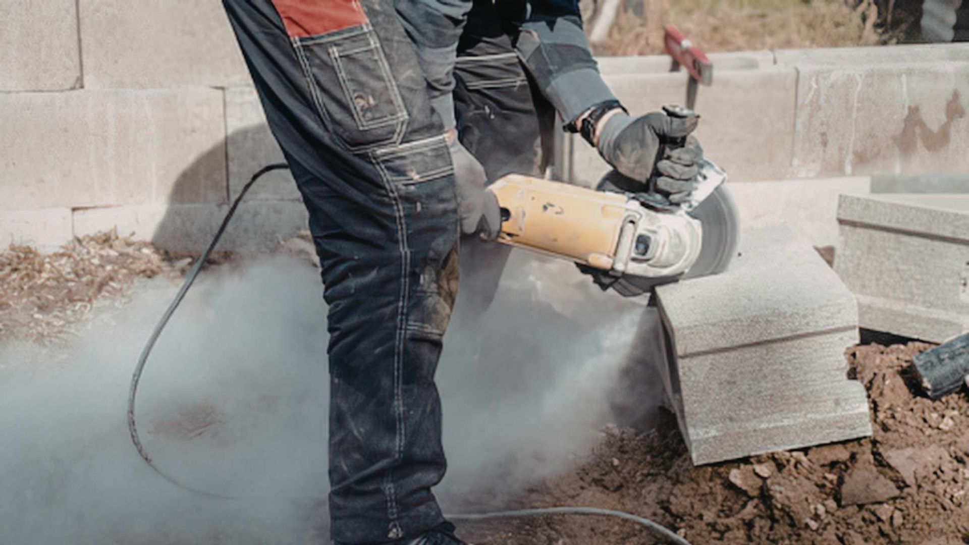 A person uses a handheld power saw to cut a concrete block, creating a large cloud of dust at a construction site.