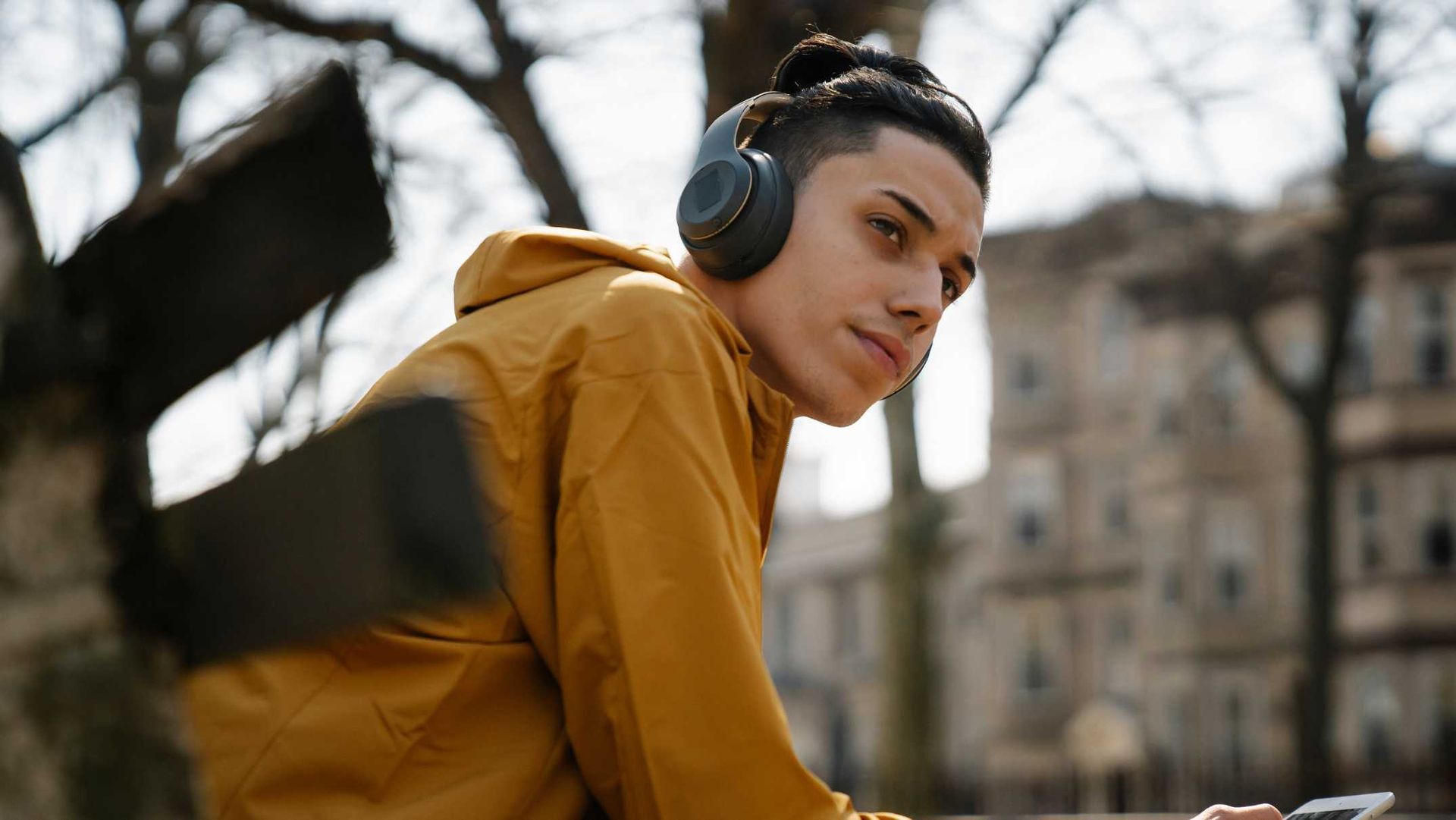 A person wearing headphones and a yellow jacket looks to the side in an outdoor, city setting.
