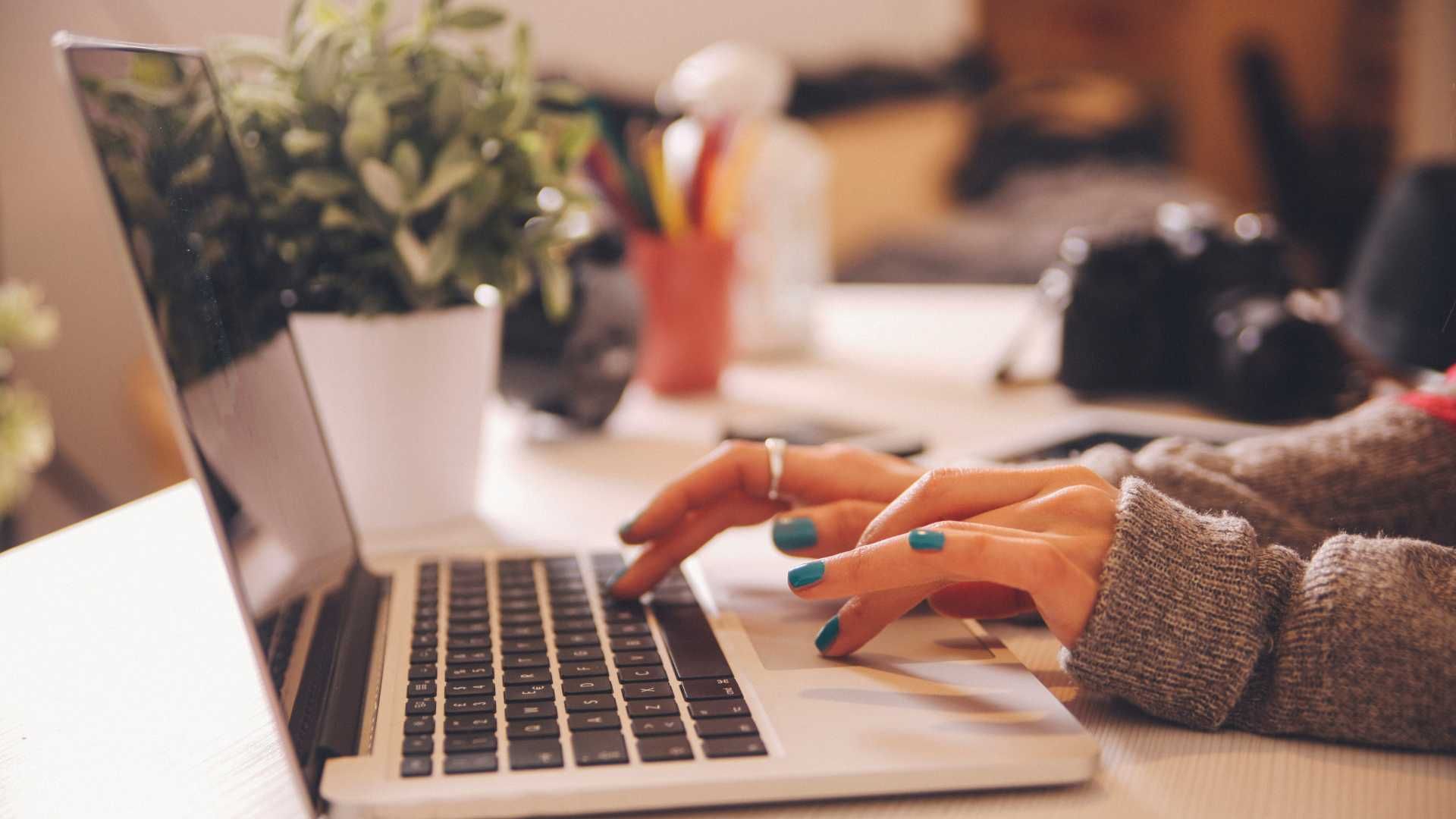 Hands with teal nail polish typing on a laptop at a desk with a plant and camera in the background.