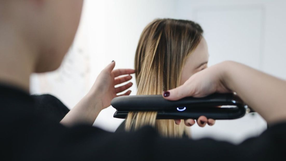 En un ambiente luminoso y minimalista, una estilista utiliza una plancha de pelo negra sobre el cabello rubio de una persona.