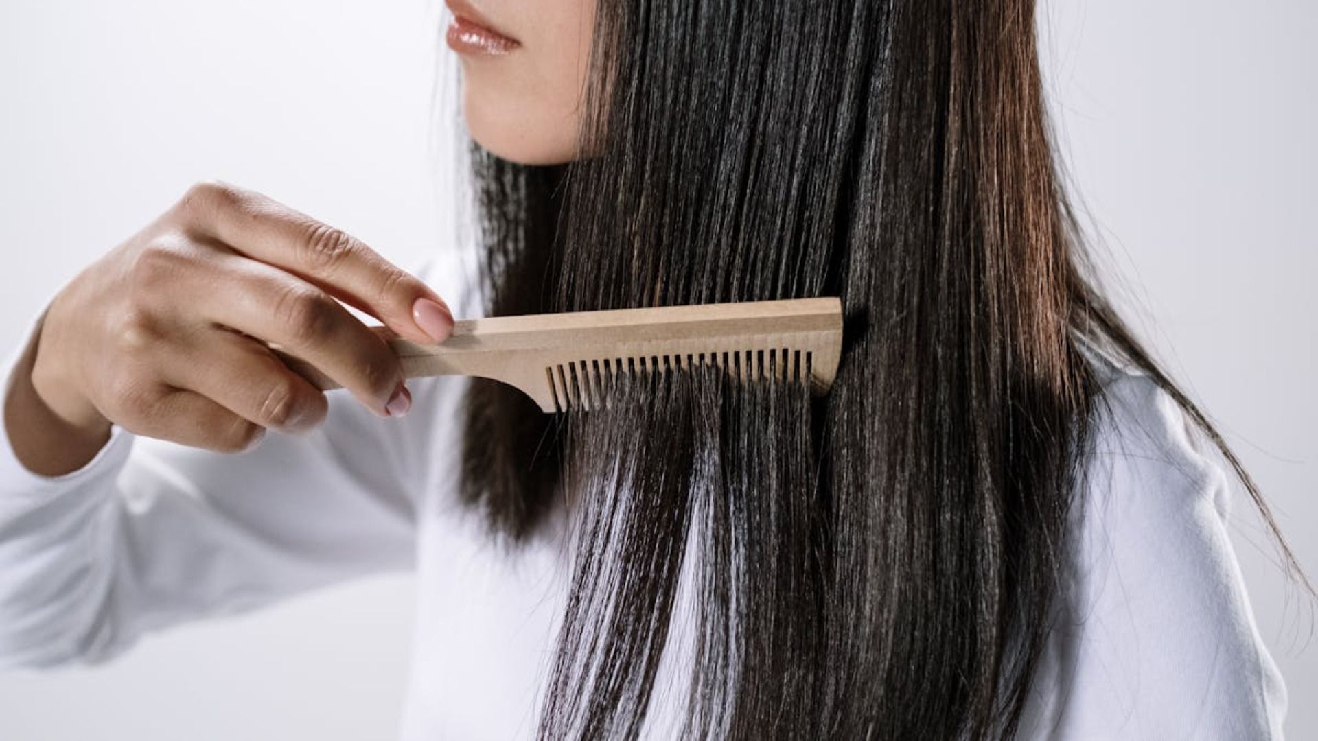Una persona con cabello largo y oscuro usa un peine de madera para alisar secciones de su cabello sobre un fondo blanco liso.