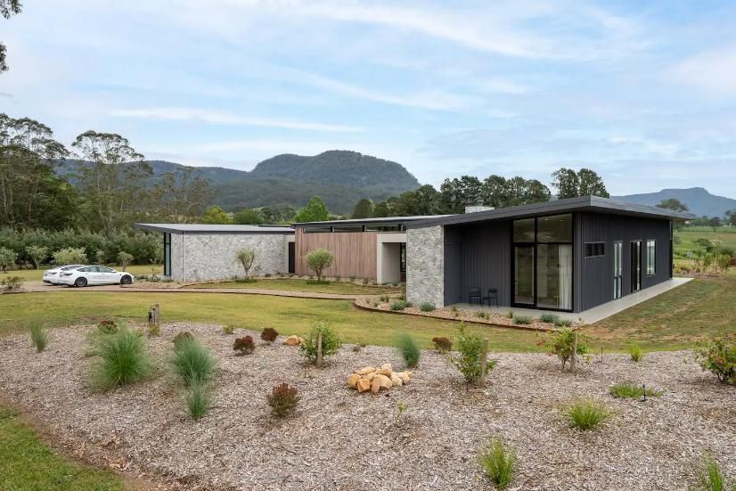 A Modern House Is Sitting In The Middle Of A Grassy Field With Mountains In The Background  — That Painter Guy In South Nowra, NSW