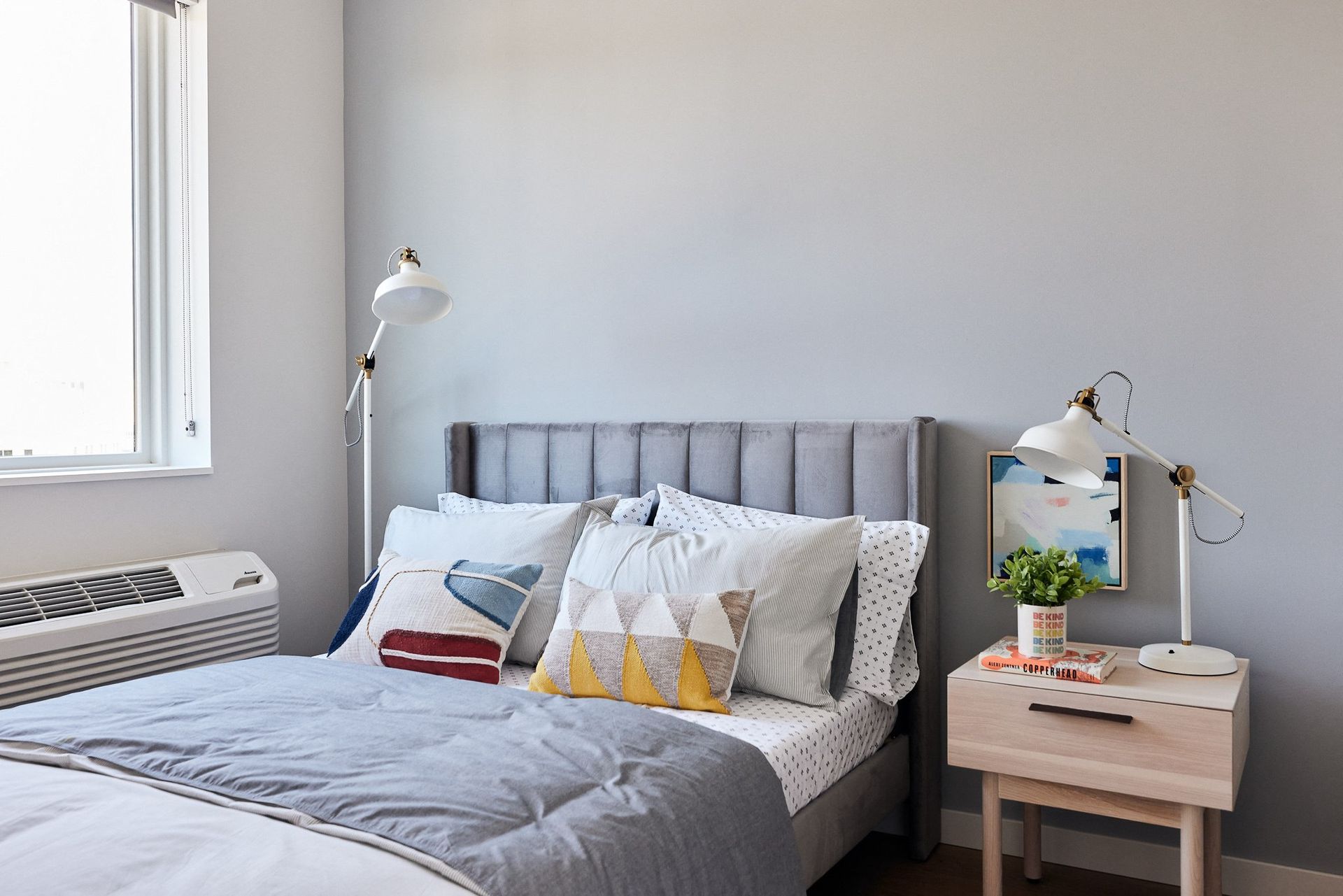 Bedroom with gray upholstered bed, light gray walls, two white desk lamps, and a wooden nightstand.