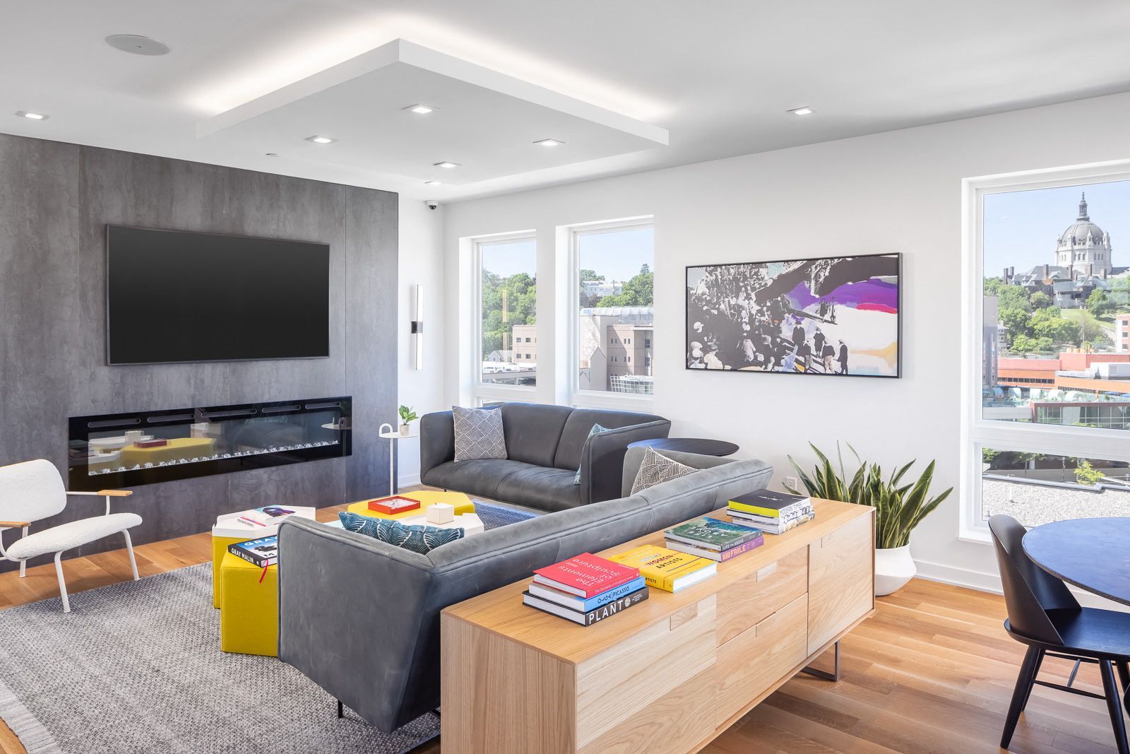 Modern living room in an apartment with a gray sectional, fireplace, wall-mounted TV, and large windows.