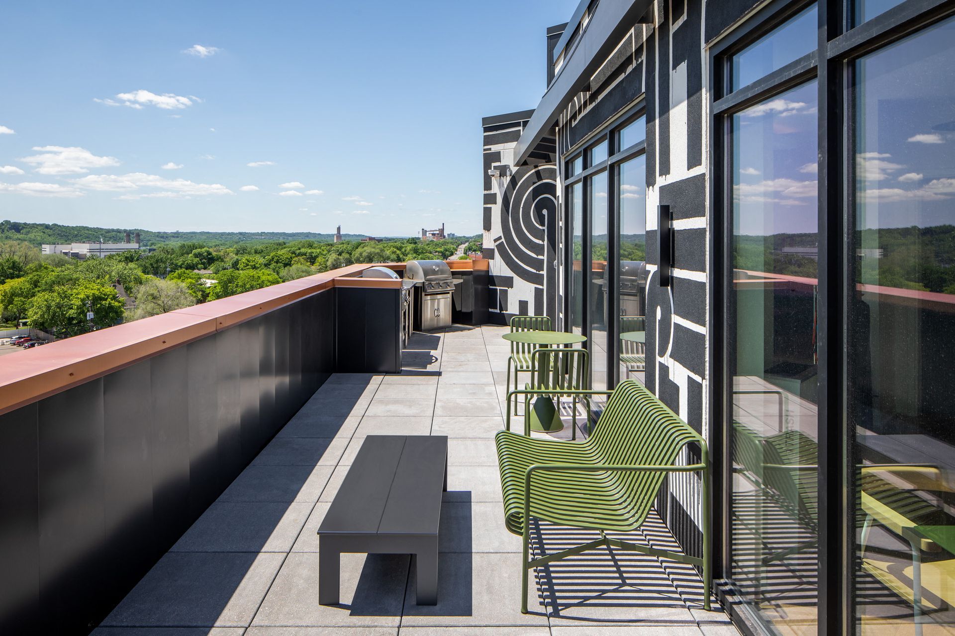 Rooftop amenity terrace with green chairs, a small table, and grill, overlooking a tree-filled landscape.