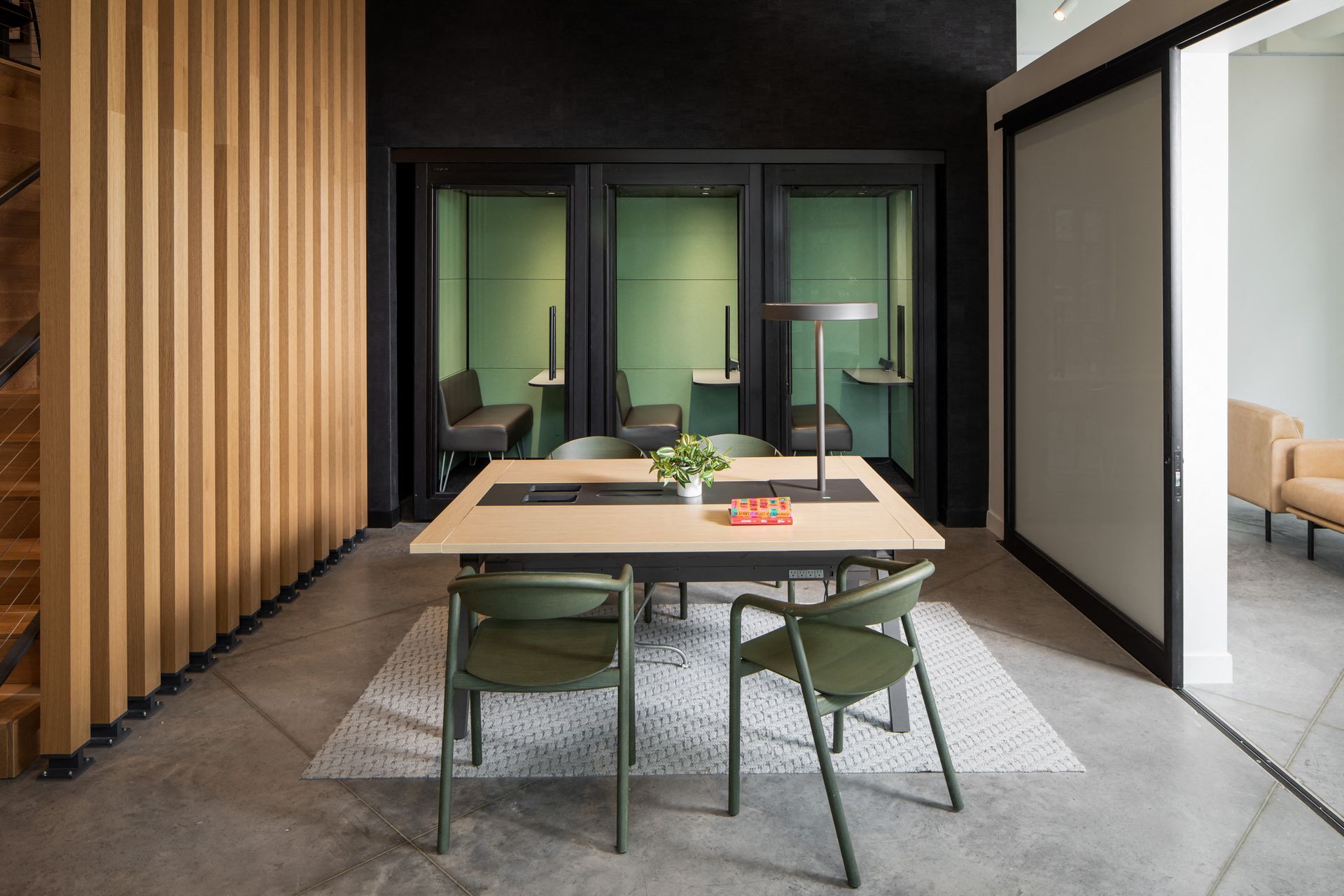 Communal meeting space with a large wooden table, green chairs, and private study pods behind glass.