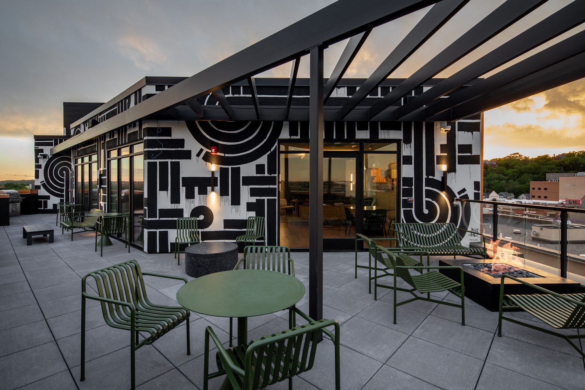Rooftop community terrace with green metal chairs, tables, and a fire pit, overlooking the city.