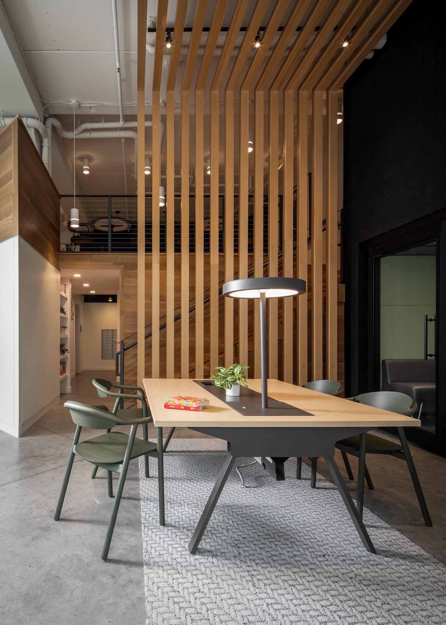 Interior communal lounge with a large wooden table, green chairs, and a vertical wood slat divider.