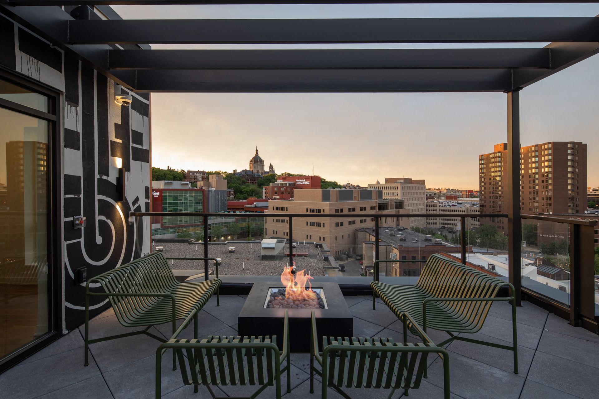 Rooftop terrace with a fire pit and green metal seating, overlooking the city skyline.