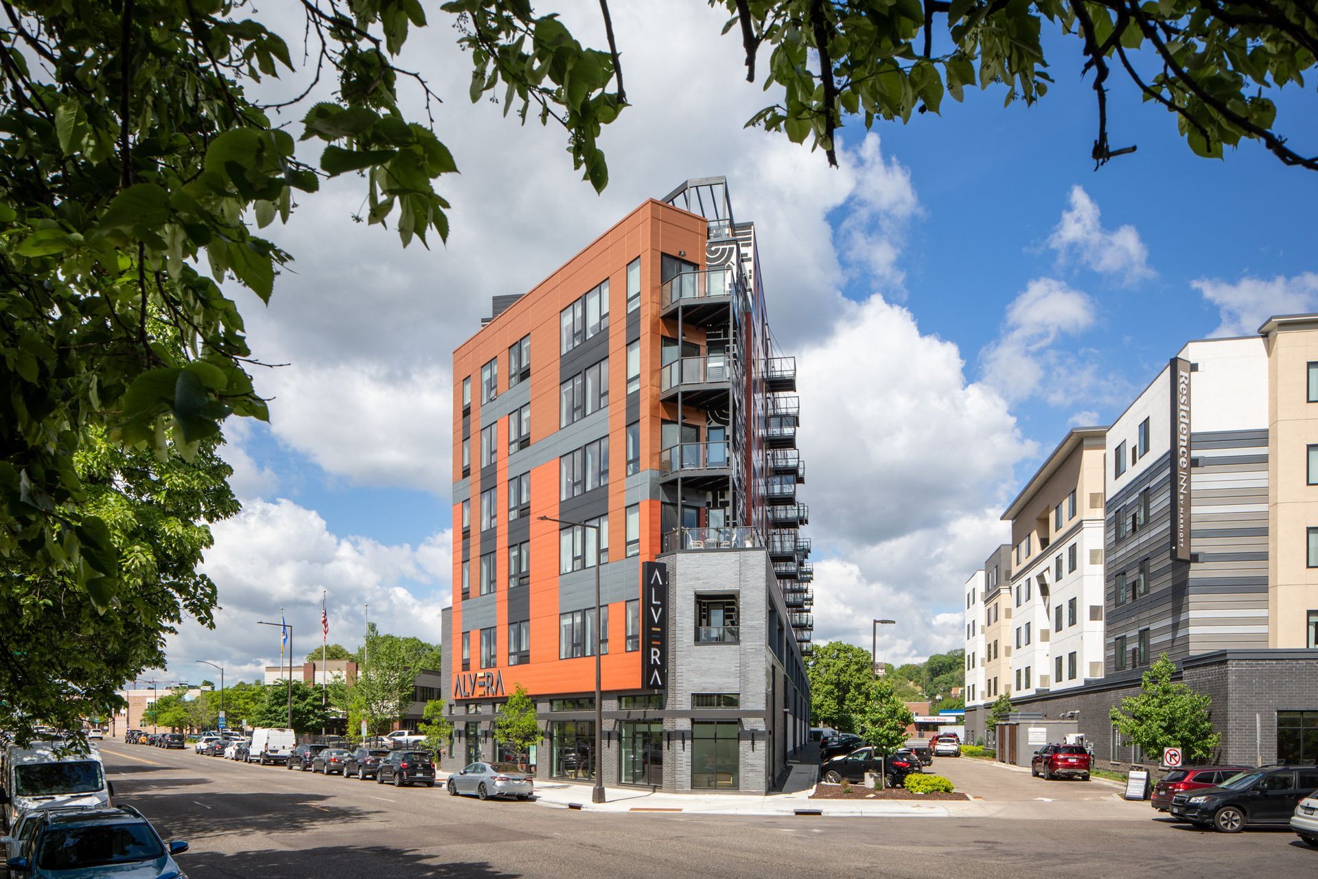 Exterior view of a modern, multi-story apartment building with orange panels and balconies on a tree-lined street.