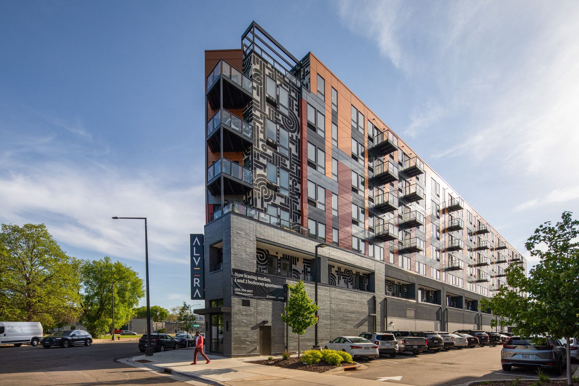 Modern, multi-story apartment building with glass balconies, colorful panels, and a street with parked cars.