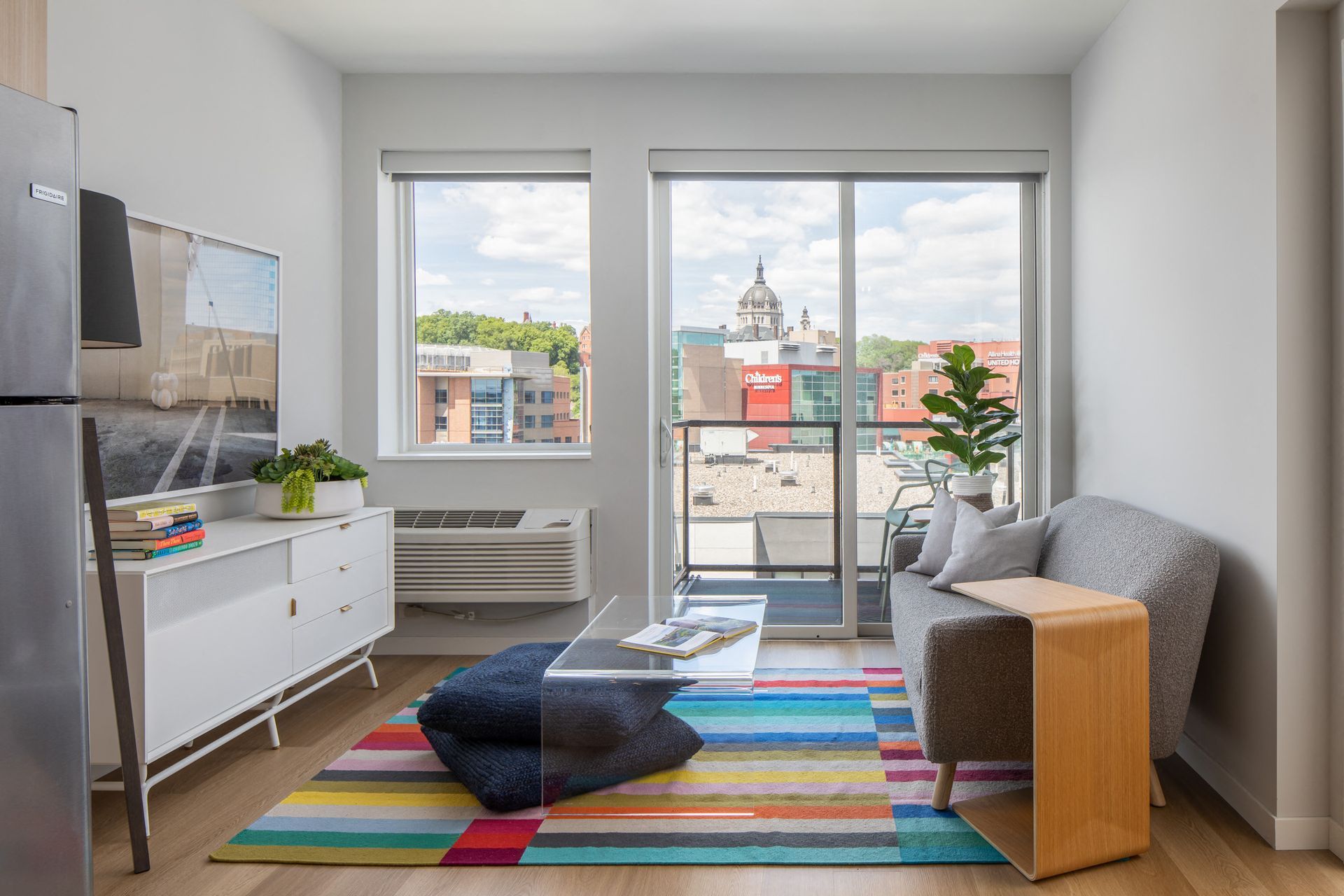 Furnished living room with sofa, glass coffee table, colorful striped rug, and balcony door with city view.