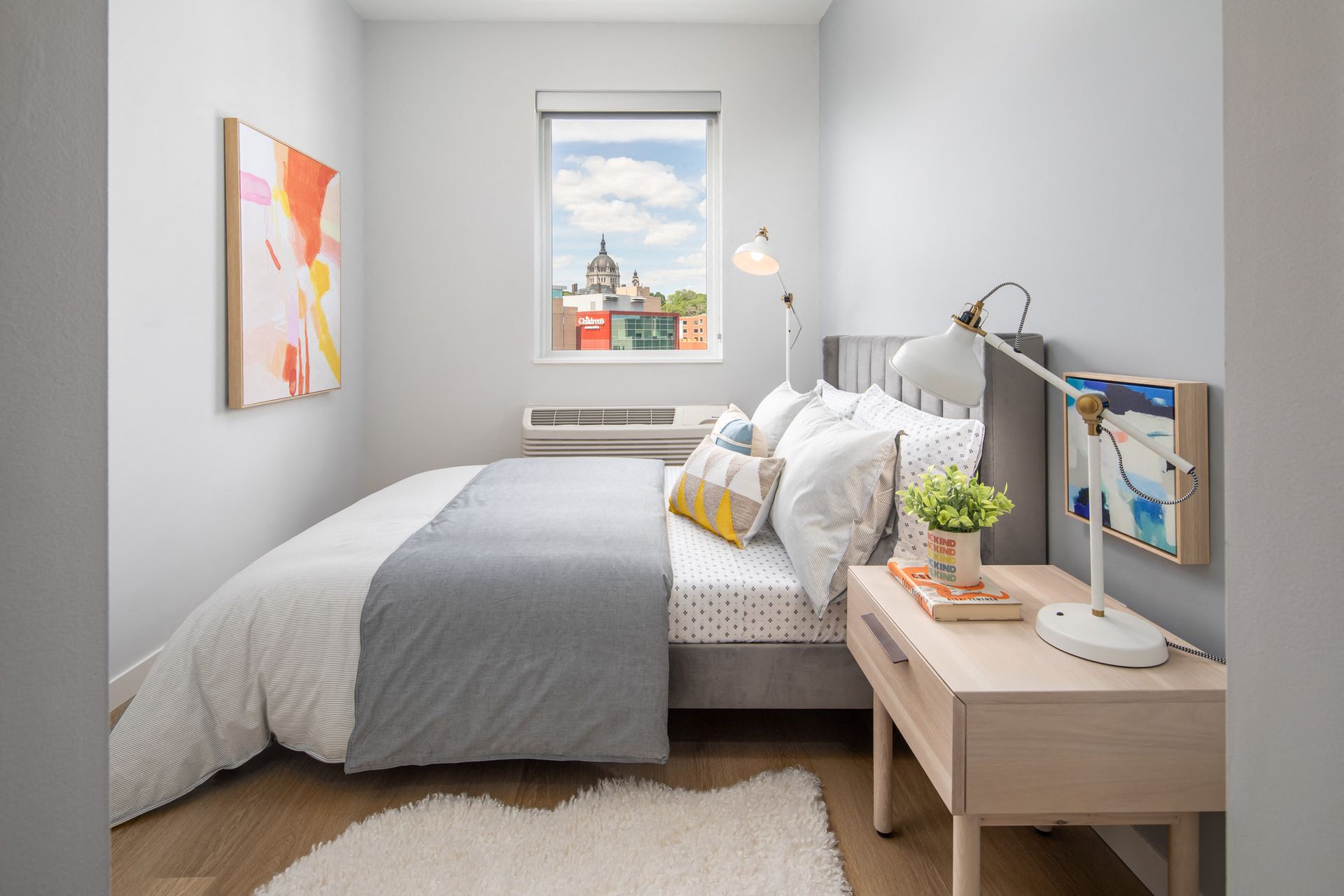 Cozy modern bedroom with a bed, nightstand, lamp, and window view of a cityscape.