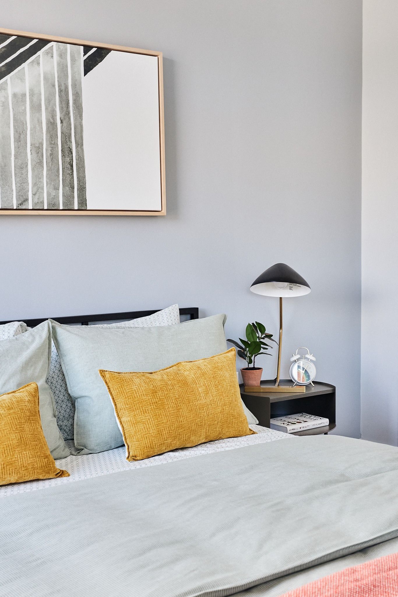 Bedroom in a modern apartment with a bed, pillows, nightstand, lamp, and a plant.