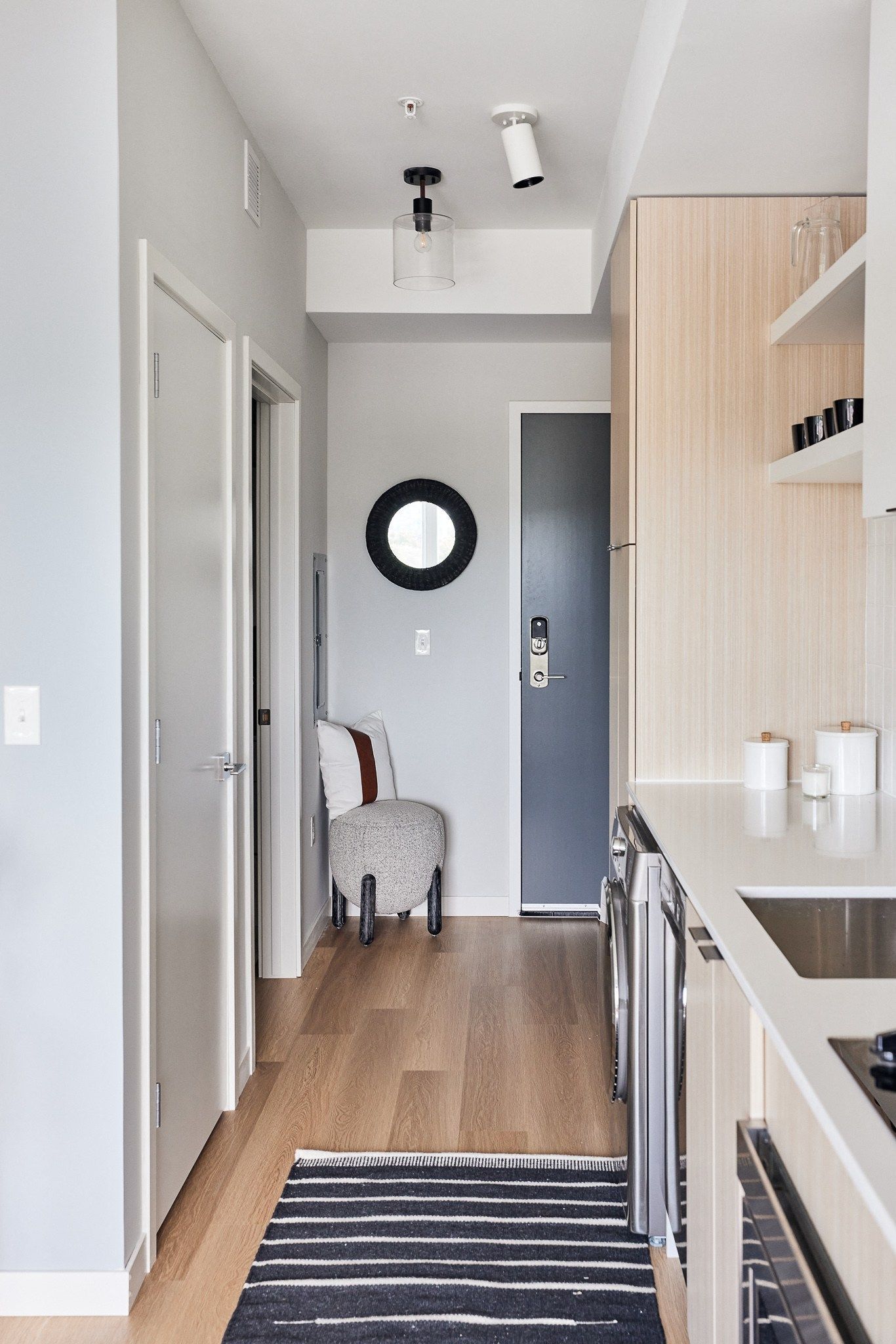 Interior view of a modern apartment kitchen and entryway with light wood cabinetry and stainless appliances.