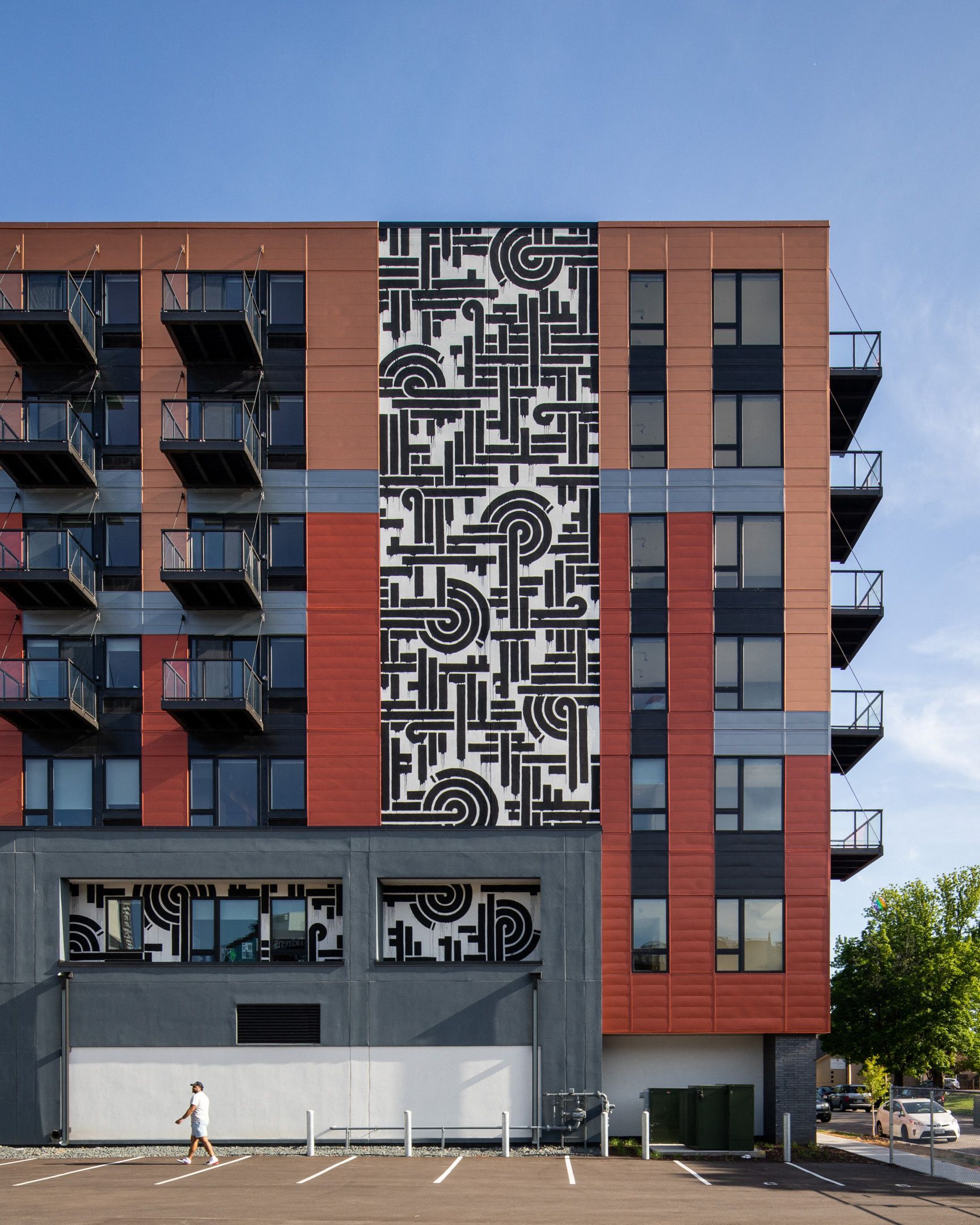 Person walking in front of a modern apartment building with red panels and a black-and-white mural.