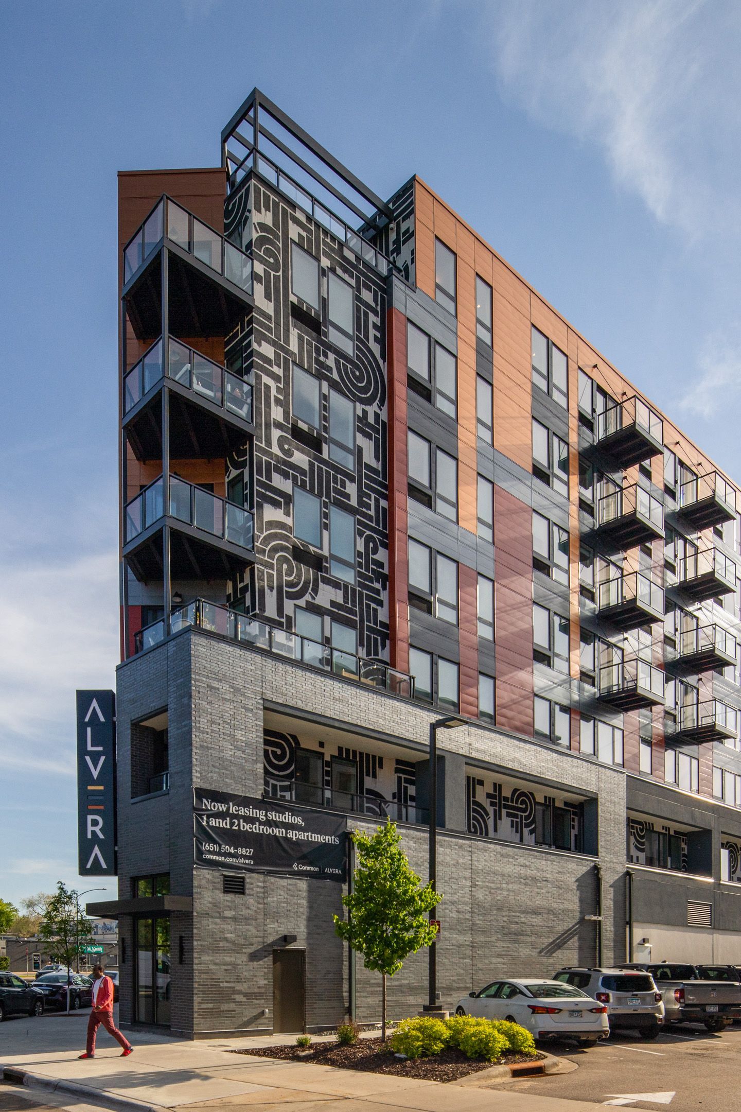 Exterior view of a modern apartment building with balconies and a mural; a person walks nearby.