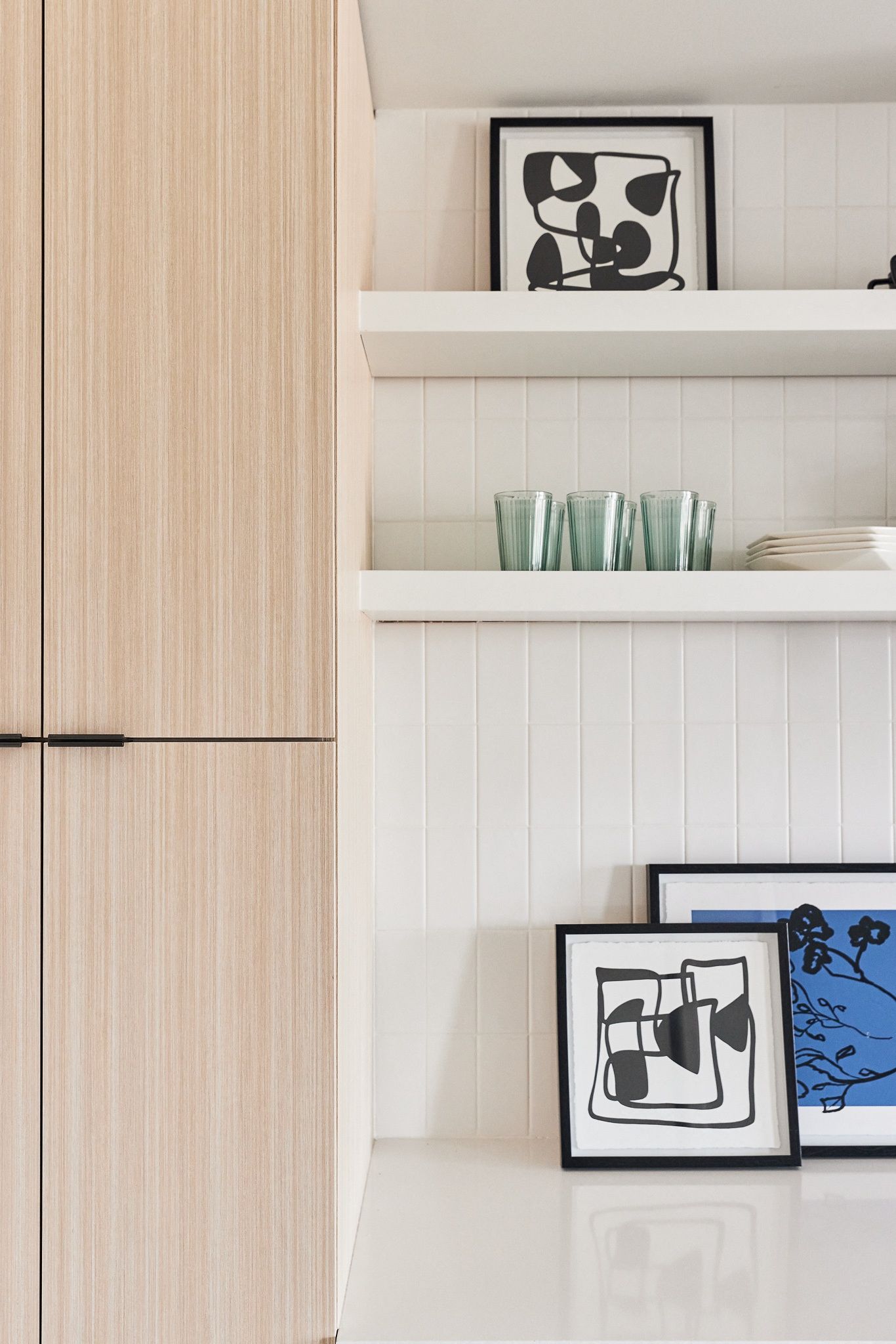 Kitchen area with light wood cabinets, white tiled backsplash, floating shelves, framed art, and green drinking glasses.