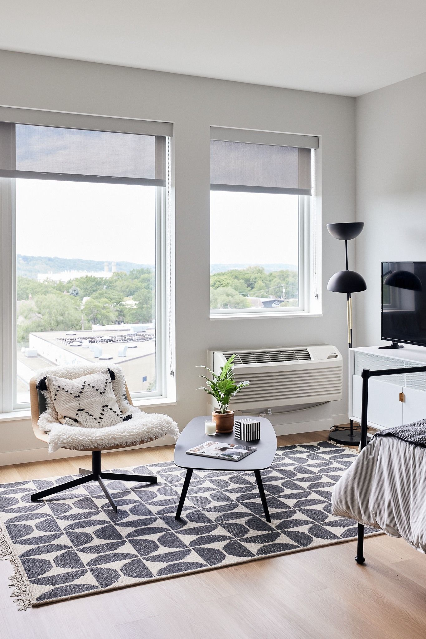 Bright apartment interior showing a bed, a chair with a sheepskin, a coffee table, rug, and large windows.