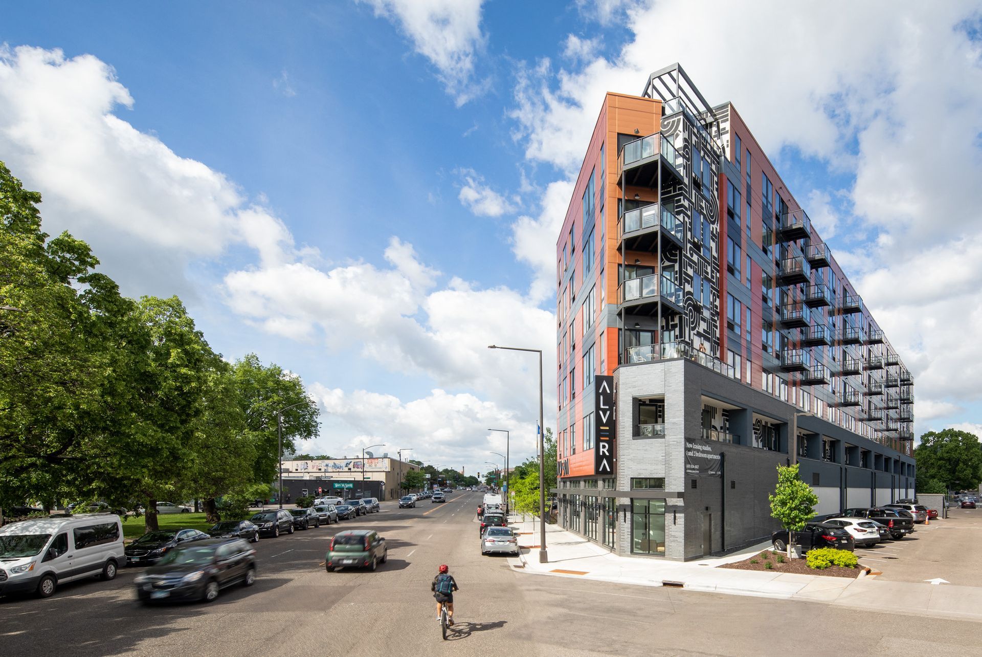 Modern mid-rise apartment building with balconies along a city street; a cyclist rides by.