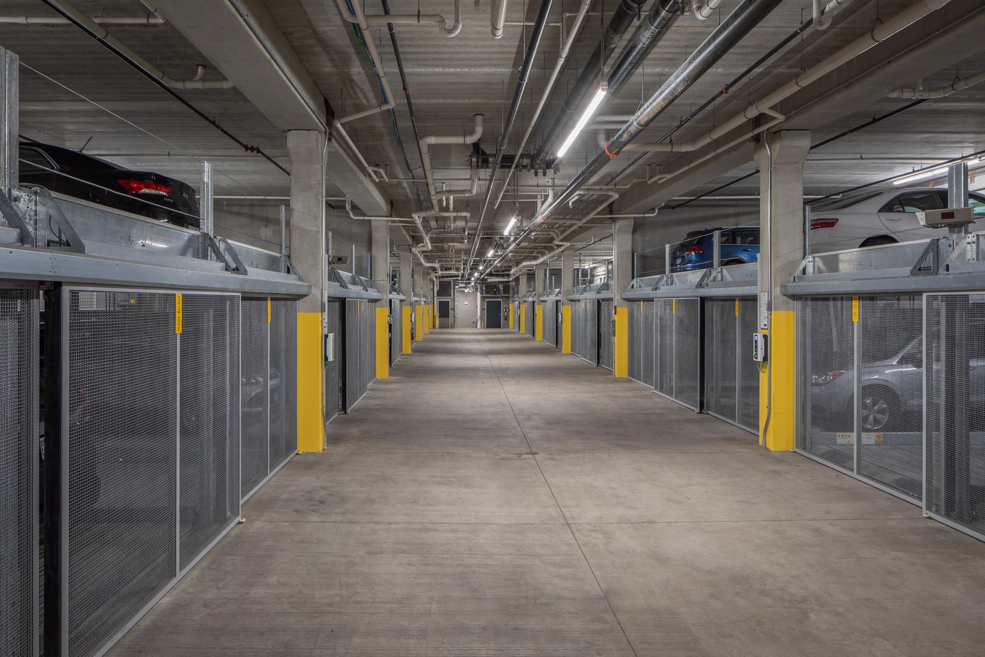 Underground parking garage with a long central aisle, mesh storage cages, and yellow column accents.