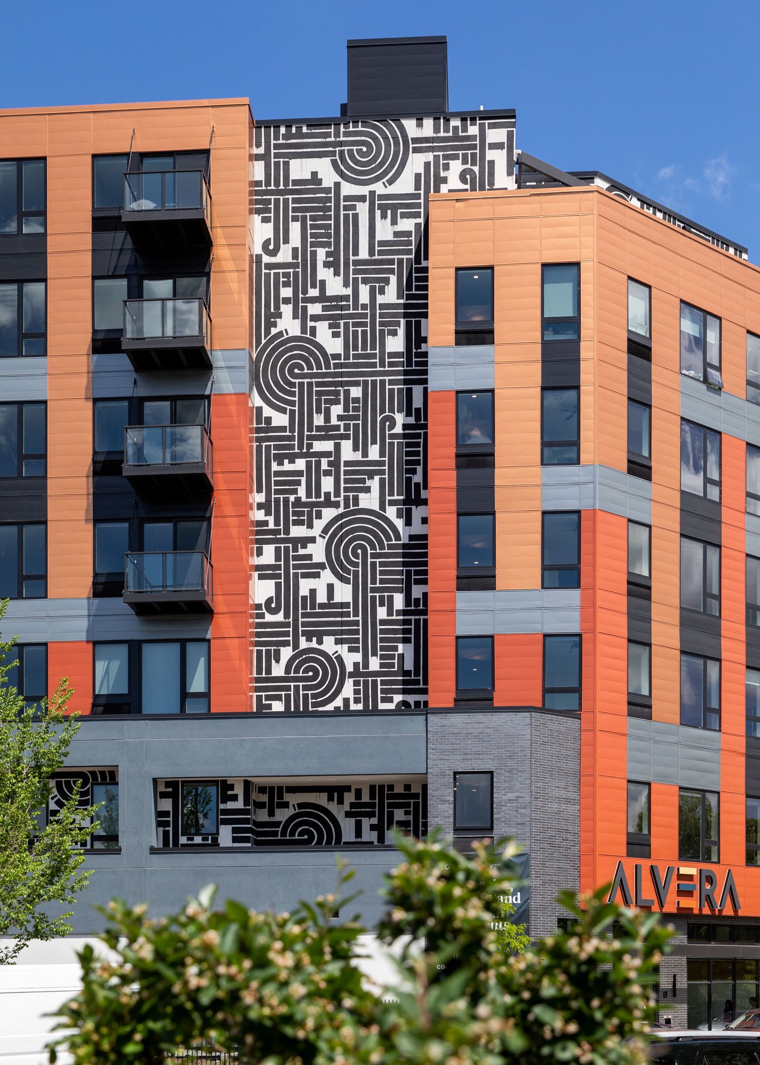 Exterior view of a modern apartment building with orange panels and a black-and-white geometric mural.