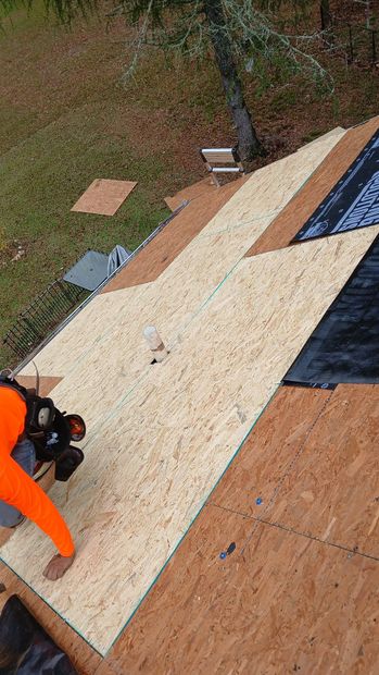 A person wearing an orange shirt works on a roof, installing wooden sheathing panels next to sections of black underlayment.