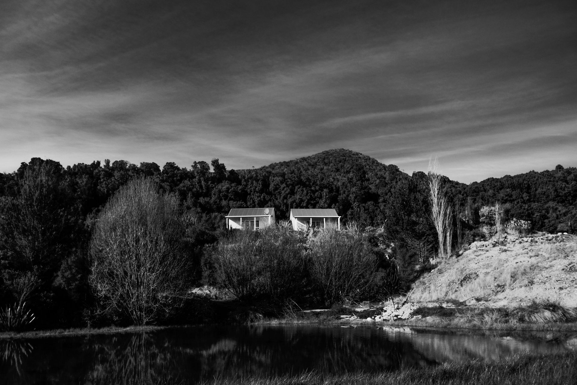 Black and white photo of two houses near a lake, surrounded by trees and a mountain, under a cloudy sky.