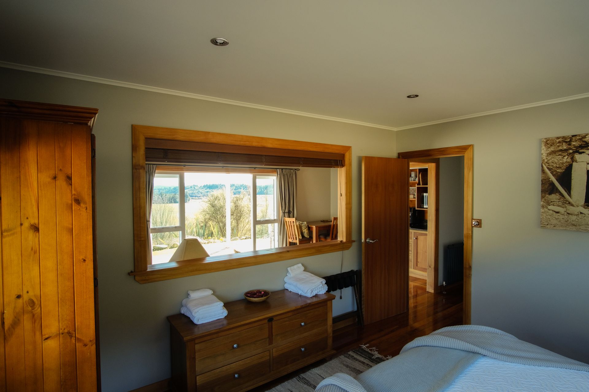Bedroom with a wooden wardrobe, dresser, and window overlooking the outdoors. A door leads to another room.