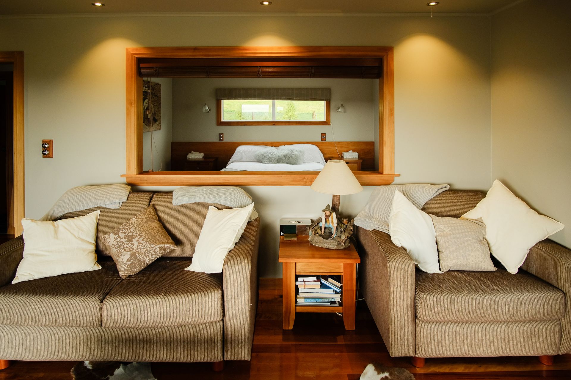 Living room with couches, a bedside table, and a window looking into a bedroom.