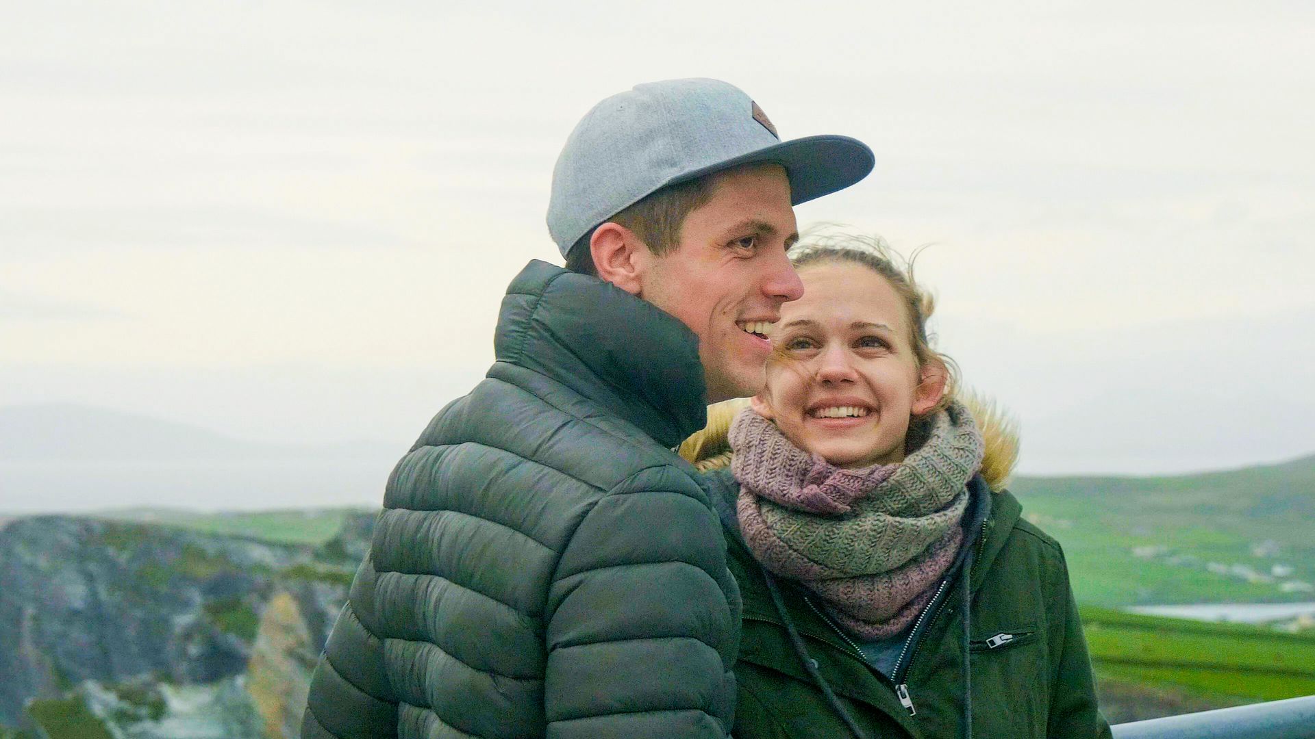 Couple smiling, looking towards a cliffside. Man wears a gray cap and puffer jacket, woman has a scarf.
