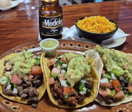 Tacos with toppings, a bowl of rice, a beer bottle, and a small cup of salsa on a wooden table.