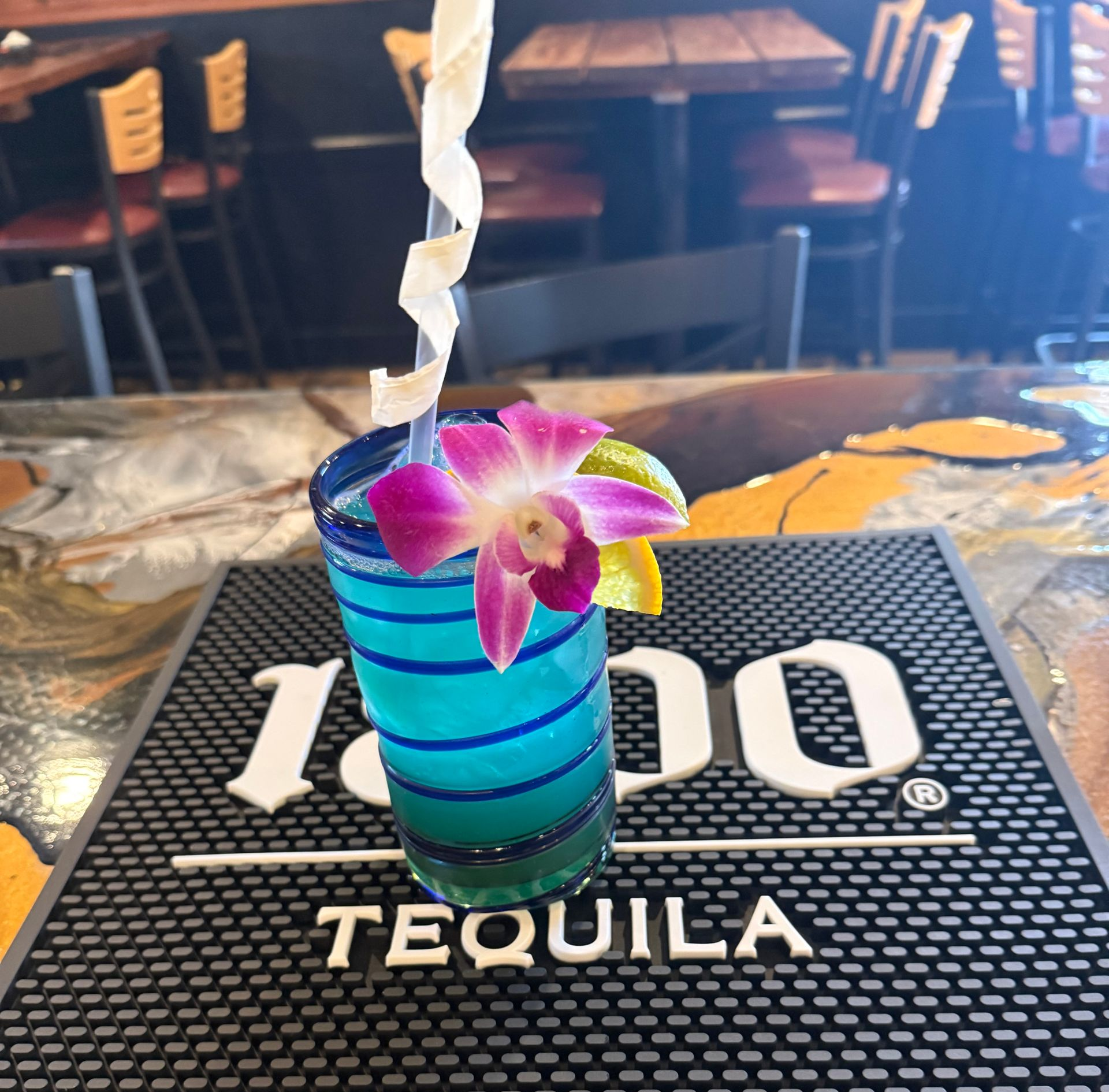 Blue cocktail with orchid garnish on a bar with a tequila logo, indoors.