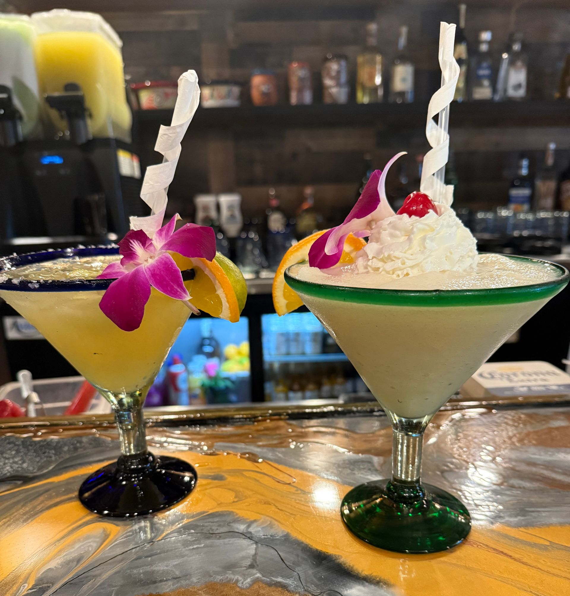 Two decorated margarita cocktails on a bar, one yellow with a flower, one white with whipped cream.