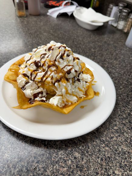 Dessert in fried pastry bowl, whipped cream, caramel, chocolate syrup, on white plate, dark countertop.