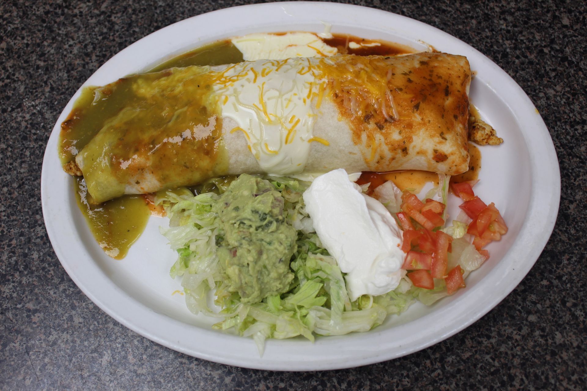 Burrito on a plate, covered in sauce, with guacamole, sour cream, and diced tomatoes.