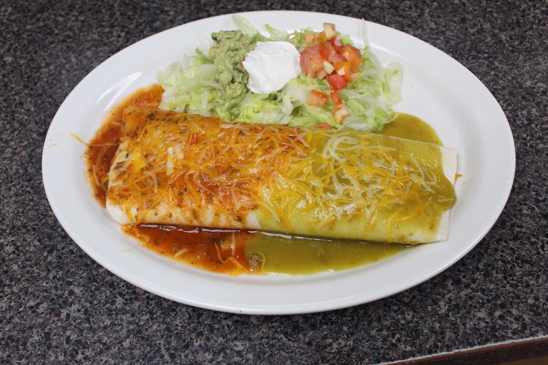 Burrito covered in red and green sauces, shredded lettuce, guacamole, sour cream, and salsa on a plate.