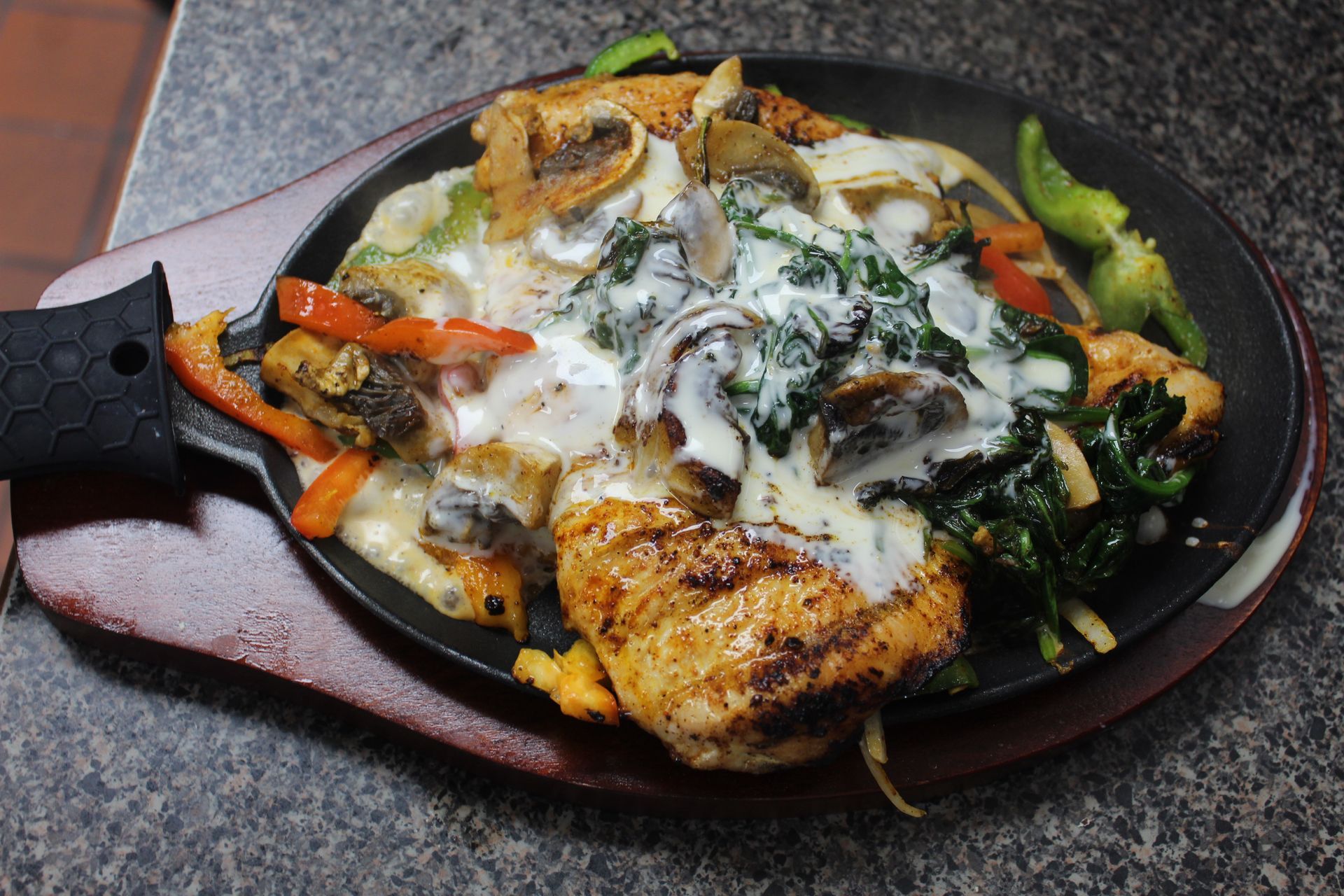Grilled chicken and vegetables with creamy sauce on a sizzling platter, served on a wooden board.