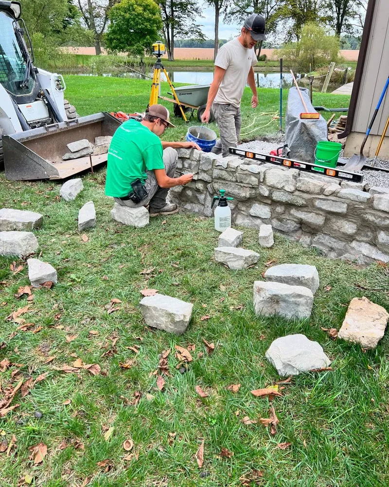 Two men are working on a stone wall in a yard.