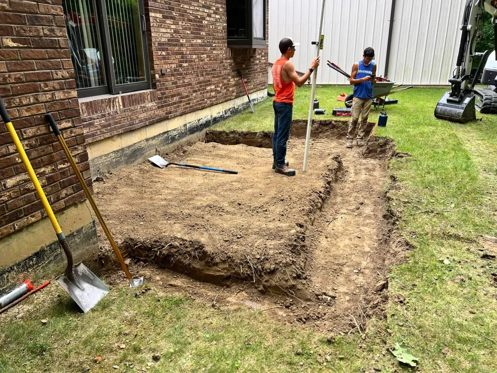 Two men are digging a hole in the ground in front of a brick building.