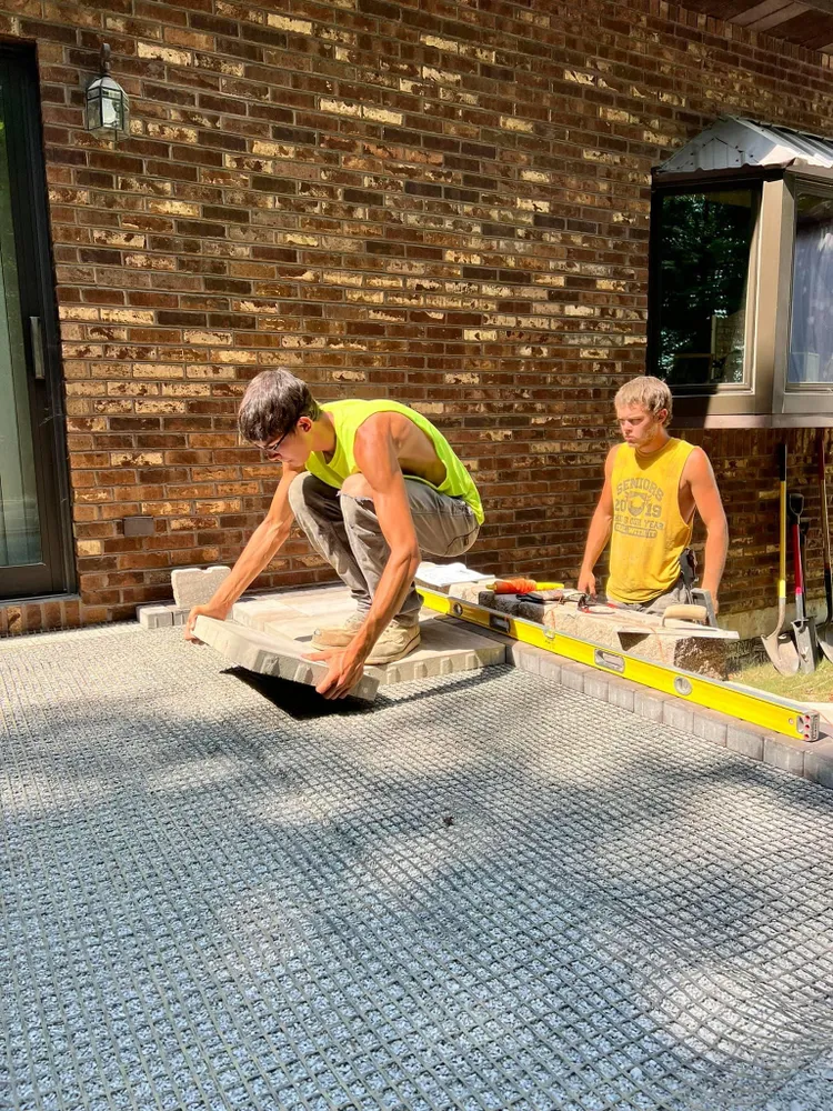 Two men are working on a patio in front of a brick building.