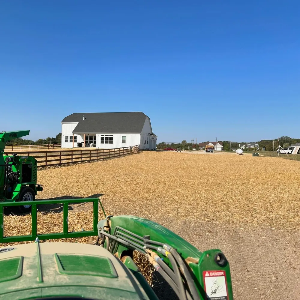 A green tractor is in a field with a house in the background
