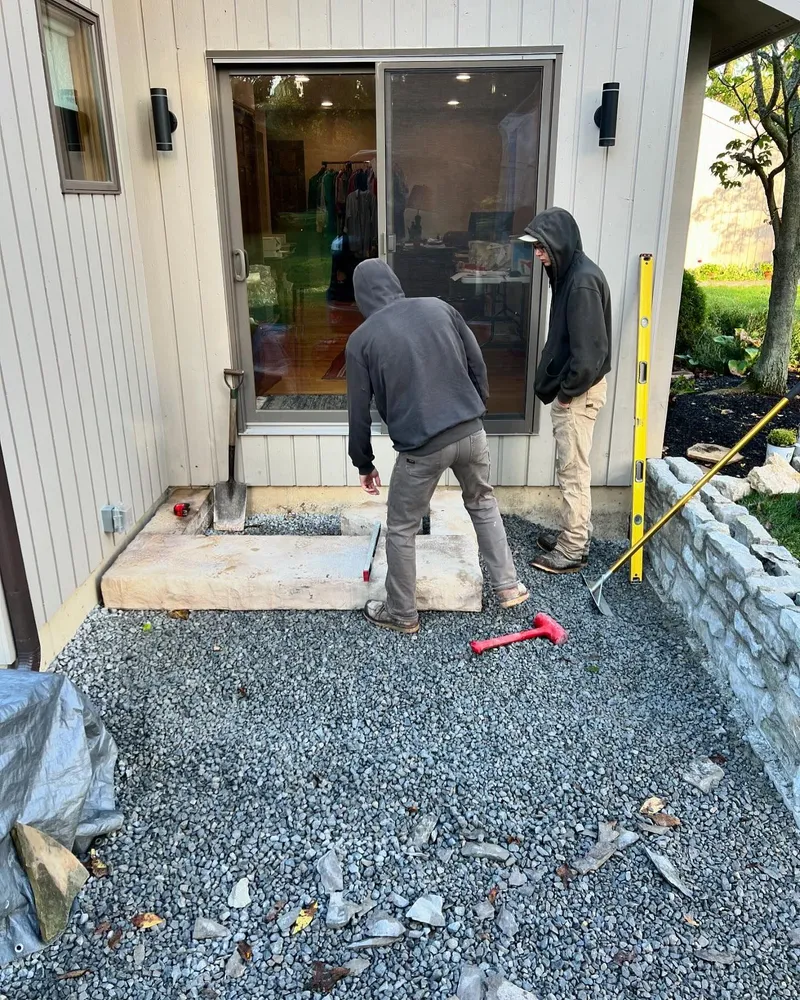 Two men are working on a patio in front of a house.