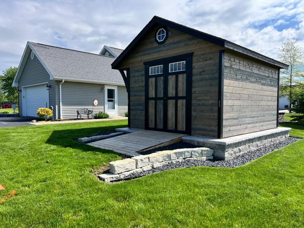 A small wooden shed is sitting in the grass in front of a house.