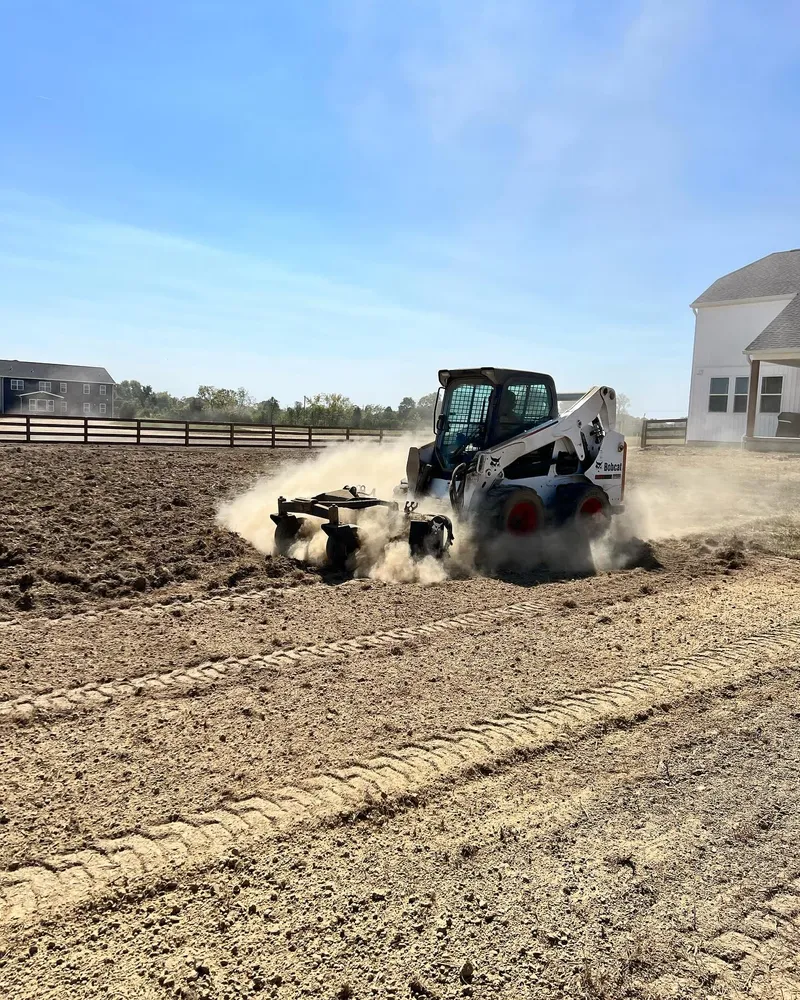 A bobcat is plowing a dirt field in front of a house.